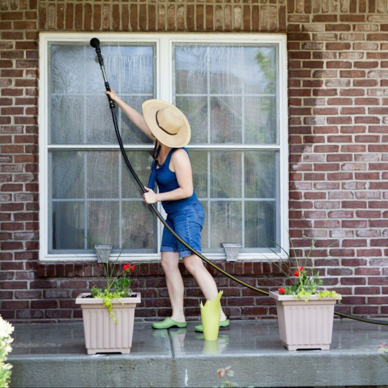 A person wearing a straw hat, blue tank top, and denim shorts uses a long hose to wash a large window on a brick house. They are standing on a wet porch next to two large planters with flowers and a green watering can.